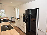 Wohnküche in Weiß und Holz mit Side-by-Side Kühlschrank, BORA-Abluftsystem und ausziehbaren Essplatz.